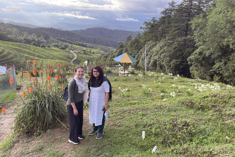 Grace Catherine Dunham and Pallavi Rajkhowa pose in front of flowers and farmland.